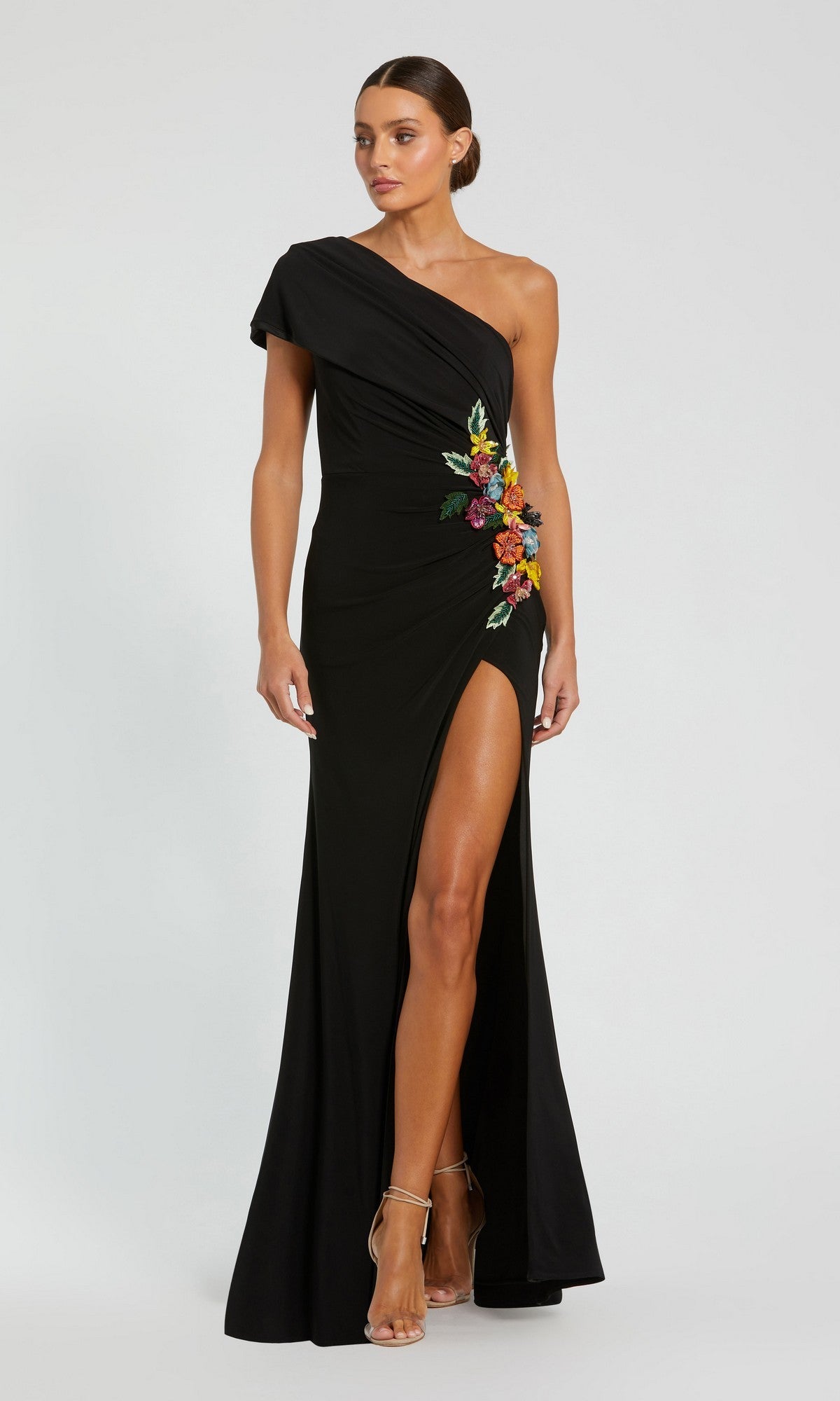 Woman wearing the Mac Duggal 11769 Long Formal Dress in black, showcasing a one-shoulder gown with a thigh-high slit and vibrant 3D floral applique embroidery at the waist, standing against a plain background.